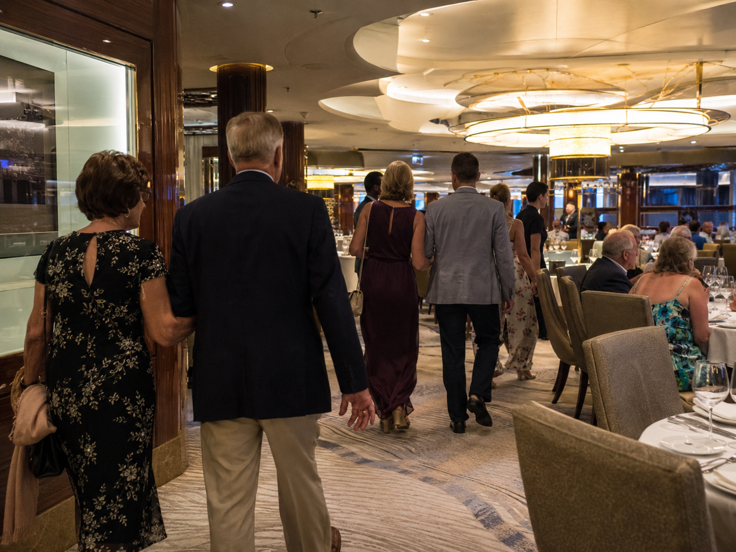 Cruise guests dressed in a mix of smart-casual and formal attire heading to dinner in a ship's main dining room